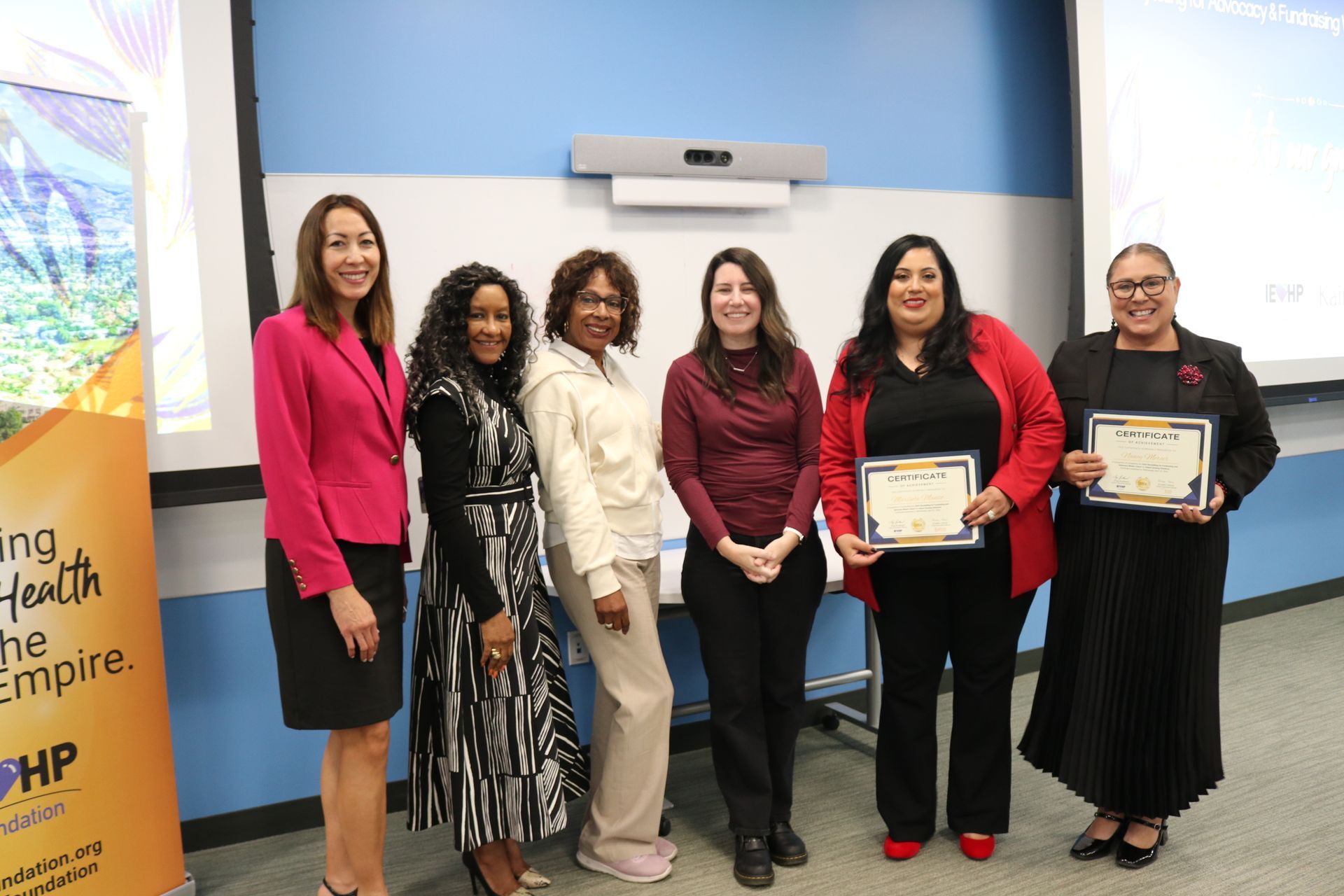 Six people posing in a conference room, two holding framed certificates in front of a projected screen.