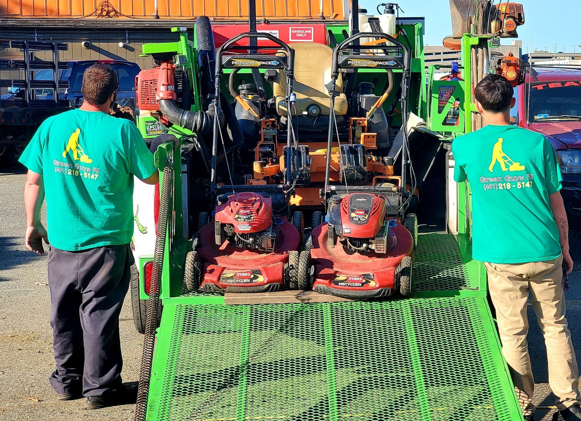 Two men in green shirts are standing in front of a trailer with lawn mowers on it.