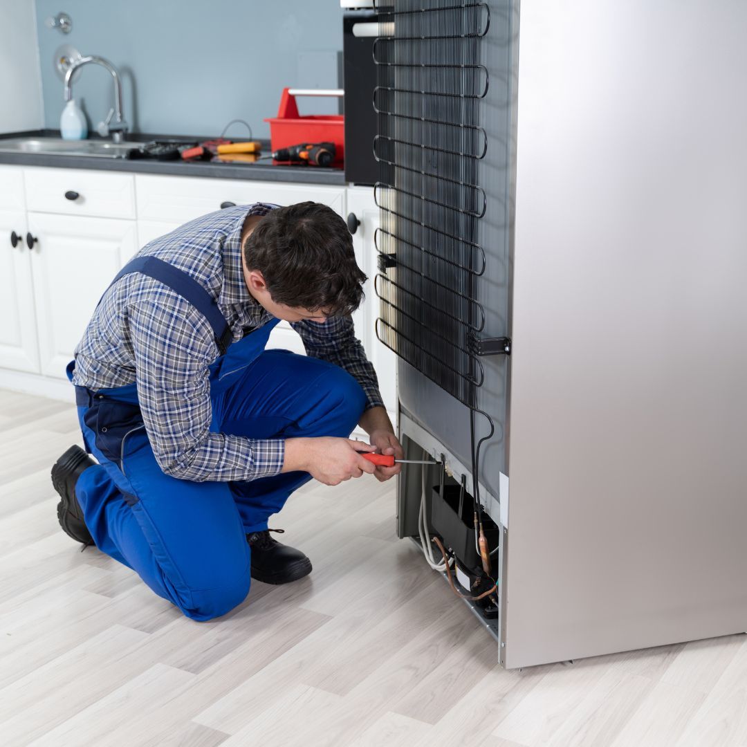 A man is kneeling down fixing a refrigerator with a screwdriver.