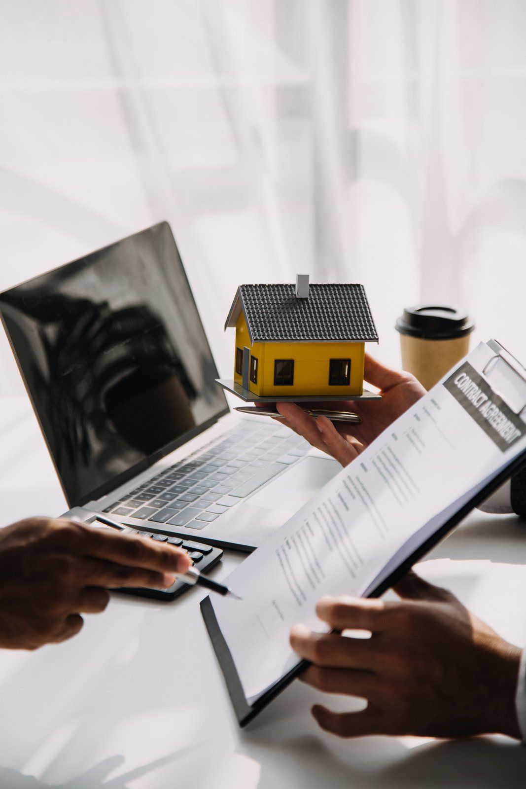 Hands holding a model house, pointing to paperwork near a laptop and coffee.