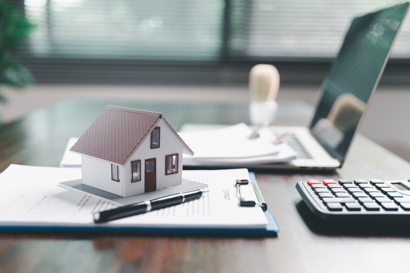 Miniature house on paperwork with pen, laptop, calculator, and stamp.