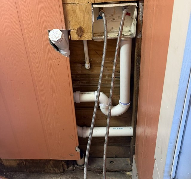 Open wall with exposed white plumbing pipes behind a broken wooden panel.