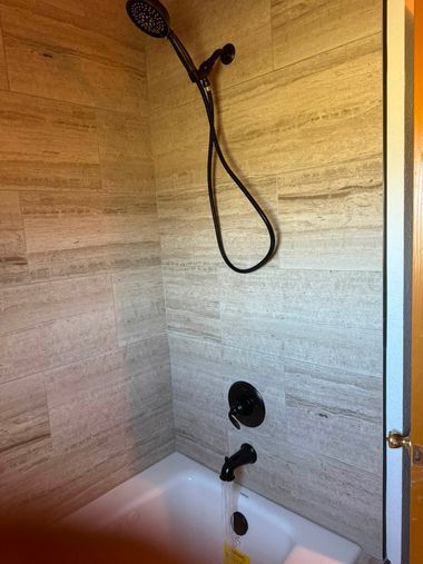 Bathroom shower with marble tile walls, handheld showerhead, and white tub beneath.