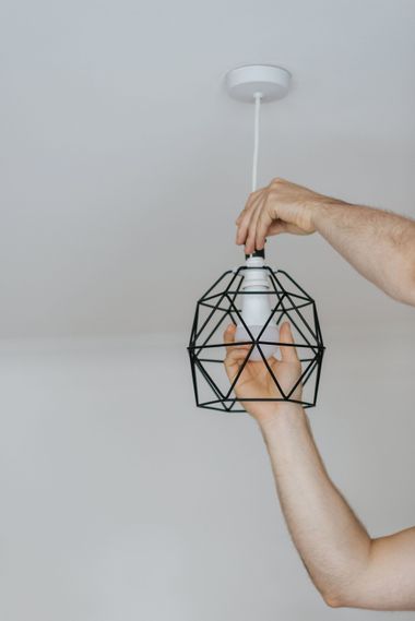 Hands changing a white light bulb in a black geometric pendant fixture on a ceiling