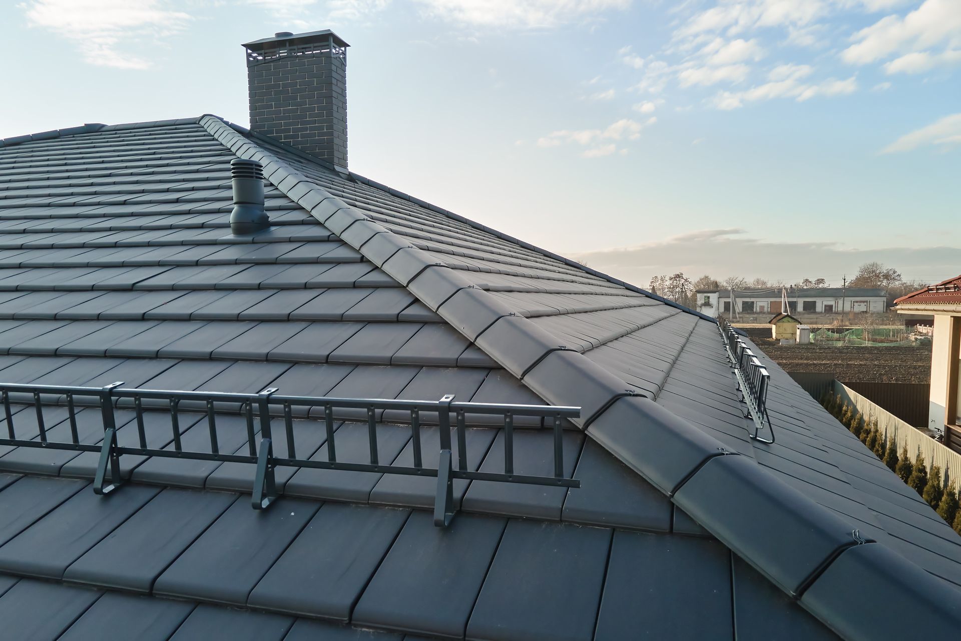 Dark Gray Tiled Roof With Safety Ladder and Chimney Against a Partly Cloudy Sky — Darren Smith Roofing In Merewether, NSW