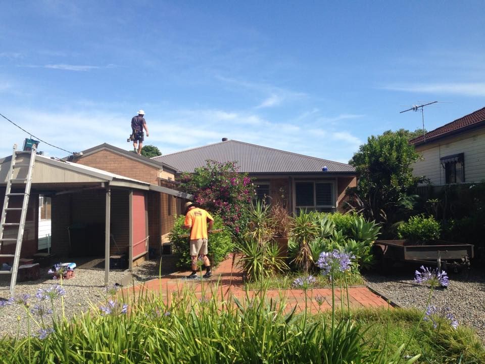 Two Workers on a Roof, One on a Shed Roof, in Front of a House With a Blue Sky — Darren Smith Roofing In Glendale, NSW
