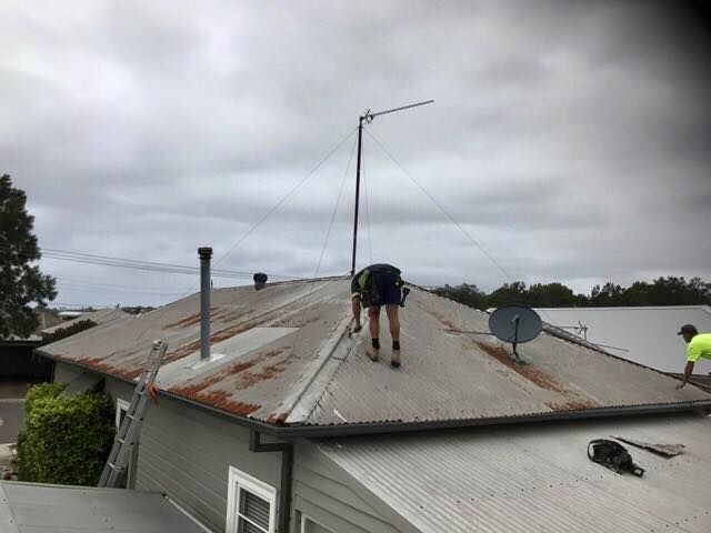 Two Workers on a Gray Corrugated Metal Roof Under a Cloudy Sky — Darren Smith Roofing In Glendale, NSW