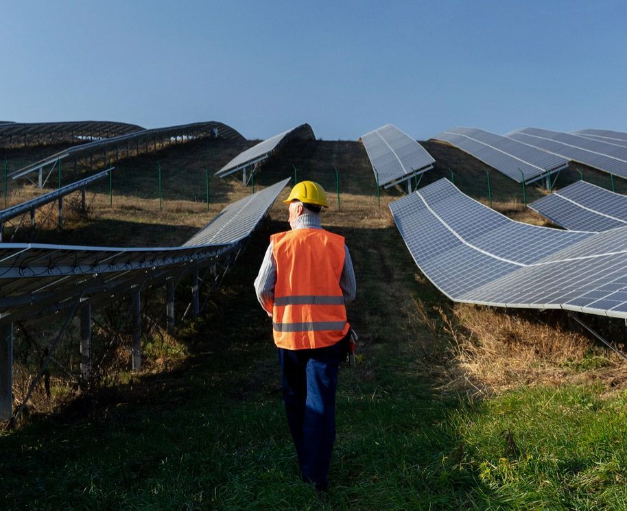 Worker in orange vest and hard hat inspecting a solar panel array on a grassy hillside.
