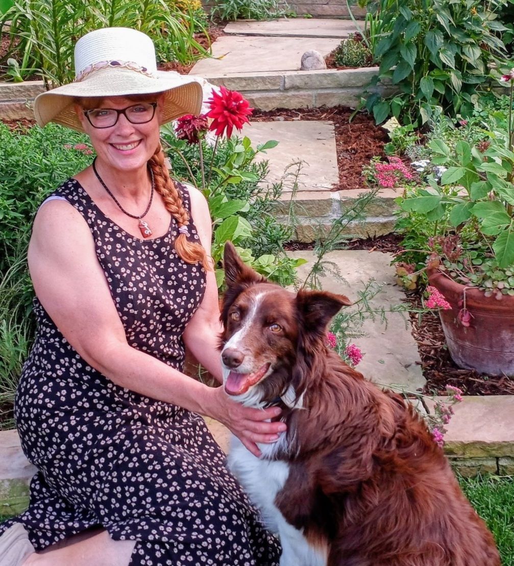Woman in hat sits with dog on stone steps in a garden.