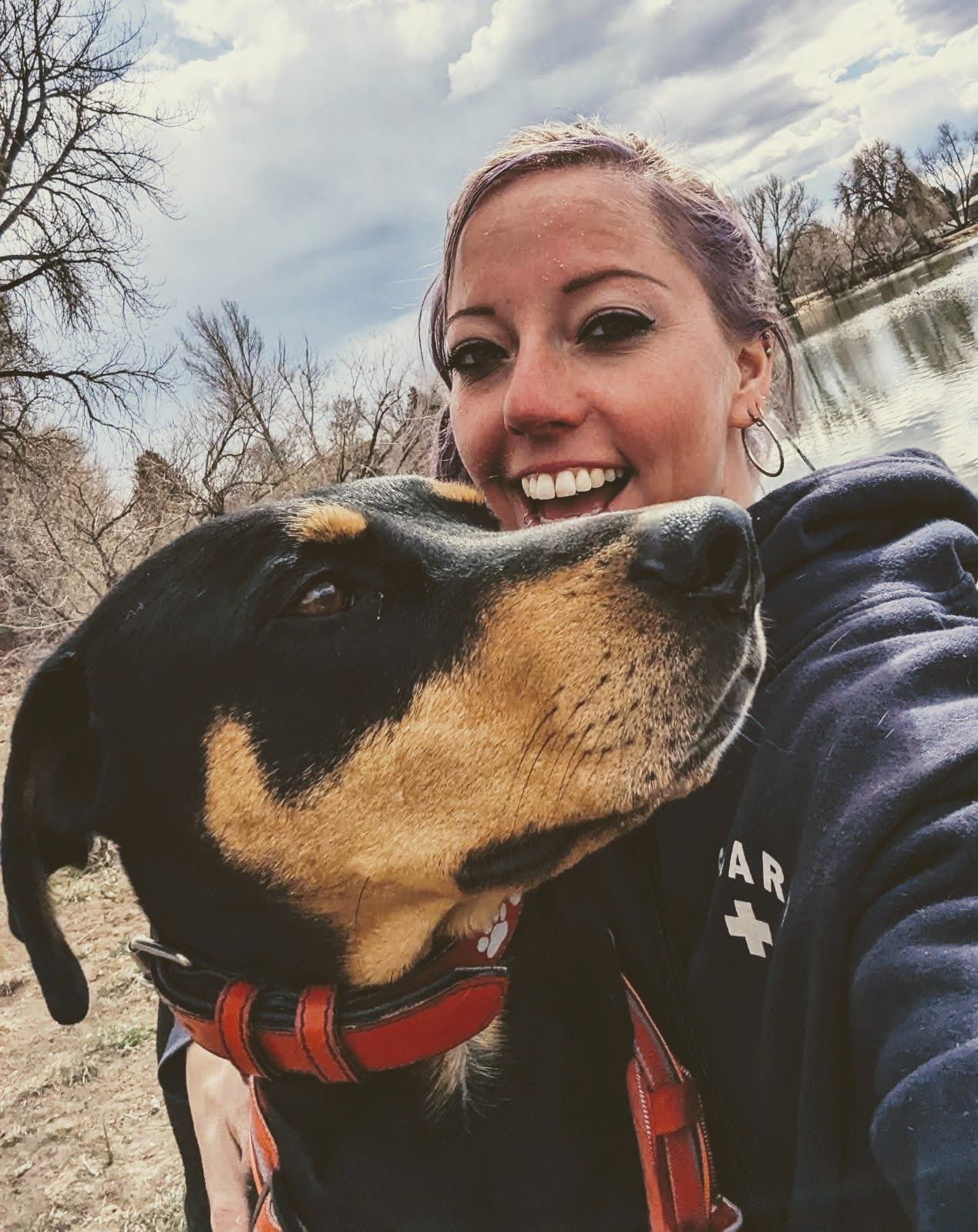 Woman smiling with a black and tan dog near a lake, both looking towards the camera.