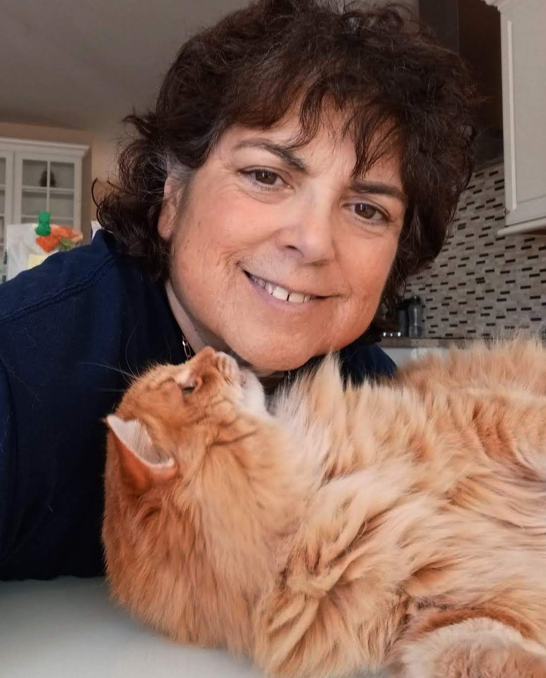 Woman with short dark hair smiles, cuddling an orange cat. Kitchen setting with white cabinets.