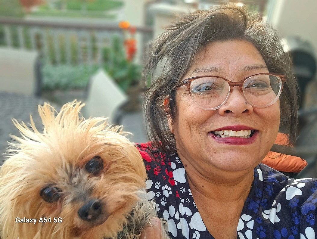 Woman smiling, holding a small brown dog outdoors. Both are looking at the camera.