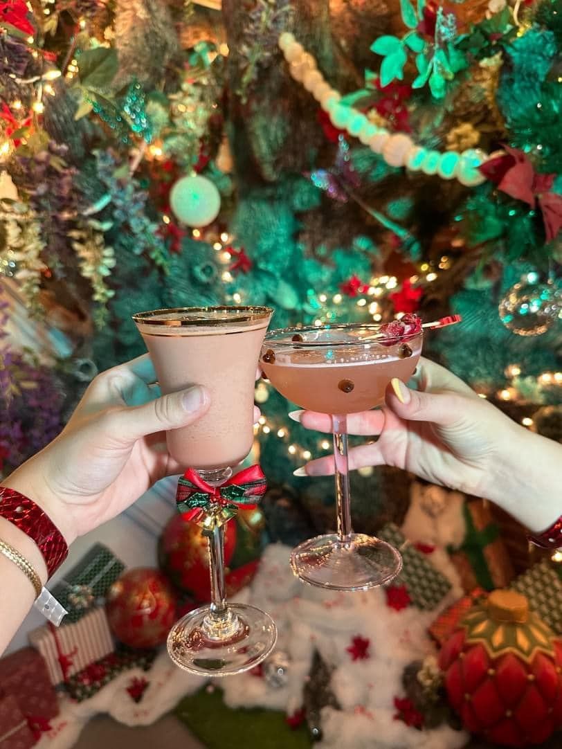 Two hands toasting cocktails in front of a decorated Christmas tree.