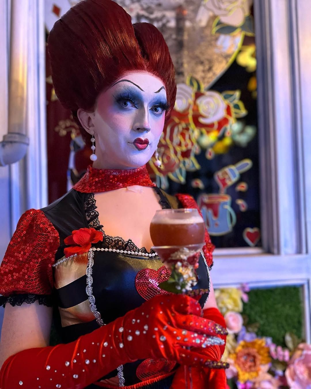 Queen of Hearts drag queen holding a drink, in red, black, and gold attire, with a decorative window backdrop.https://www.hight-orlando.com/