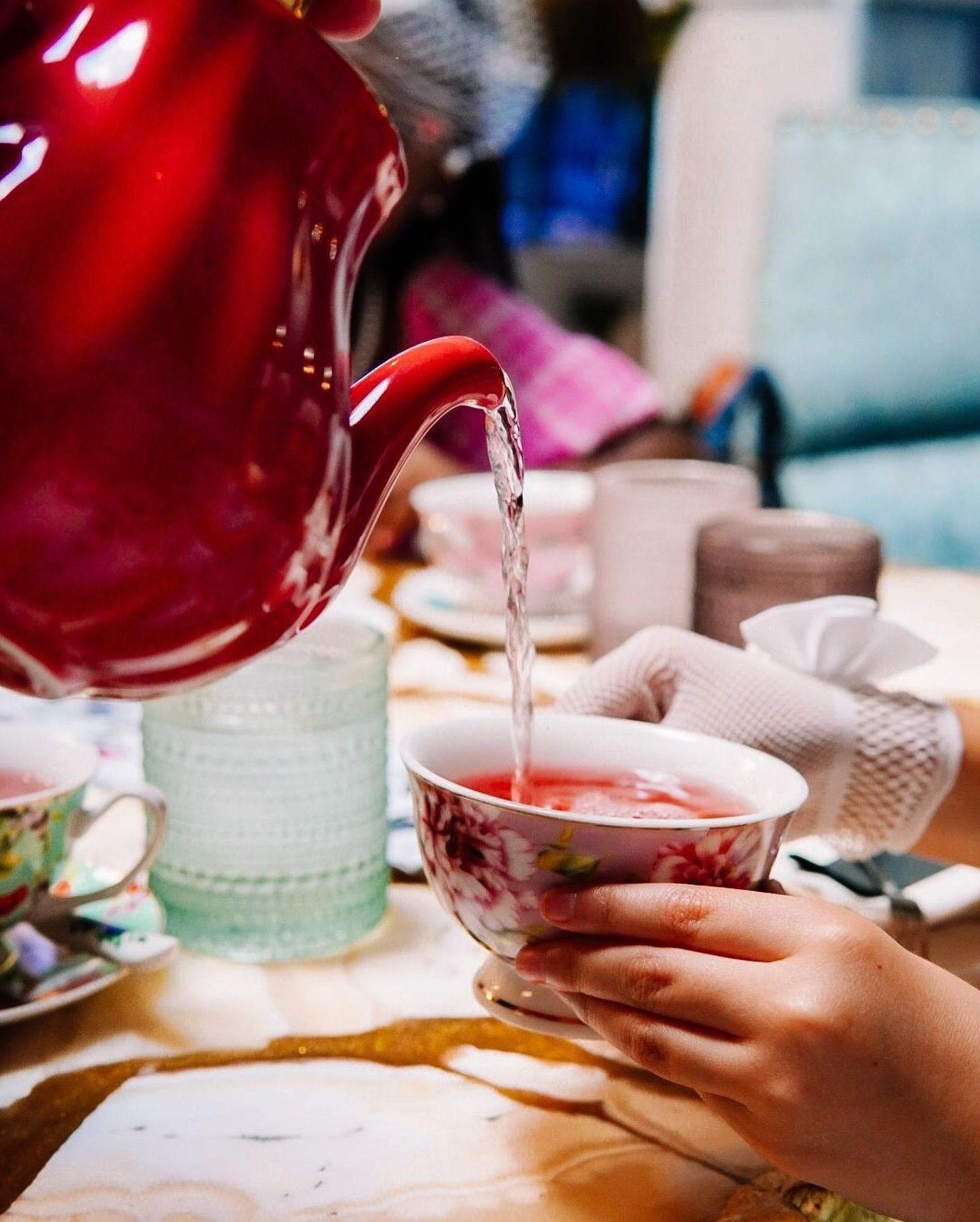 Red teapot pouring tea into floral teacup held by gloved hands at a table.https://www.hight-orlando.com/