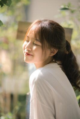 Woman smiling, eyes closed, in sunlight, wearing a white shirt; ponytail hairstyle.