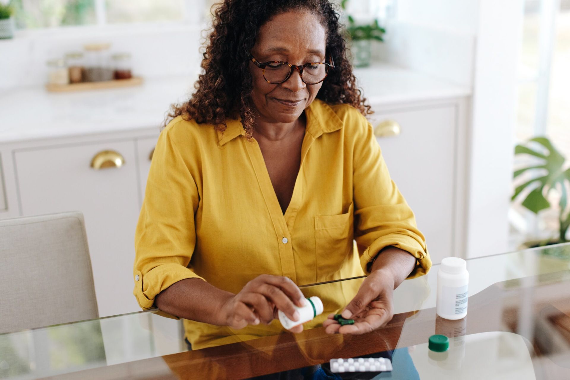 Woman pours pills from a bottle onto her hand, seated at a table. She wears a yellow shirt and glasses.
