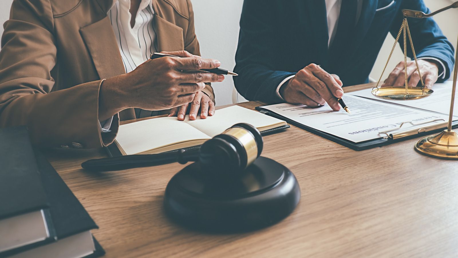 Two people in suits at a table with legal documents, scales, and a gavel.