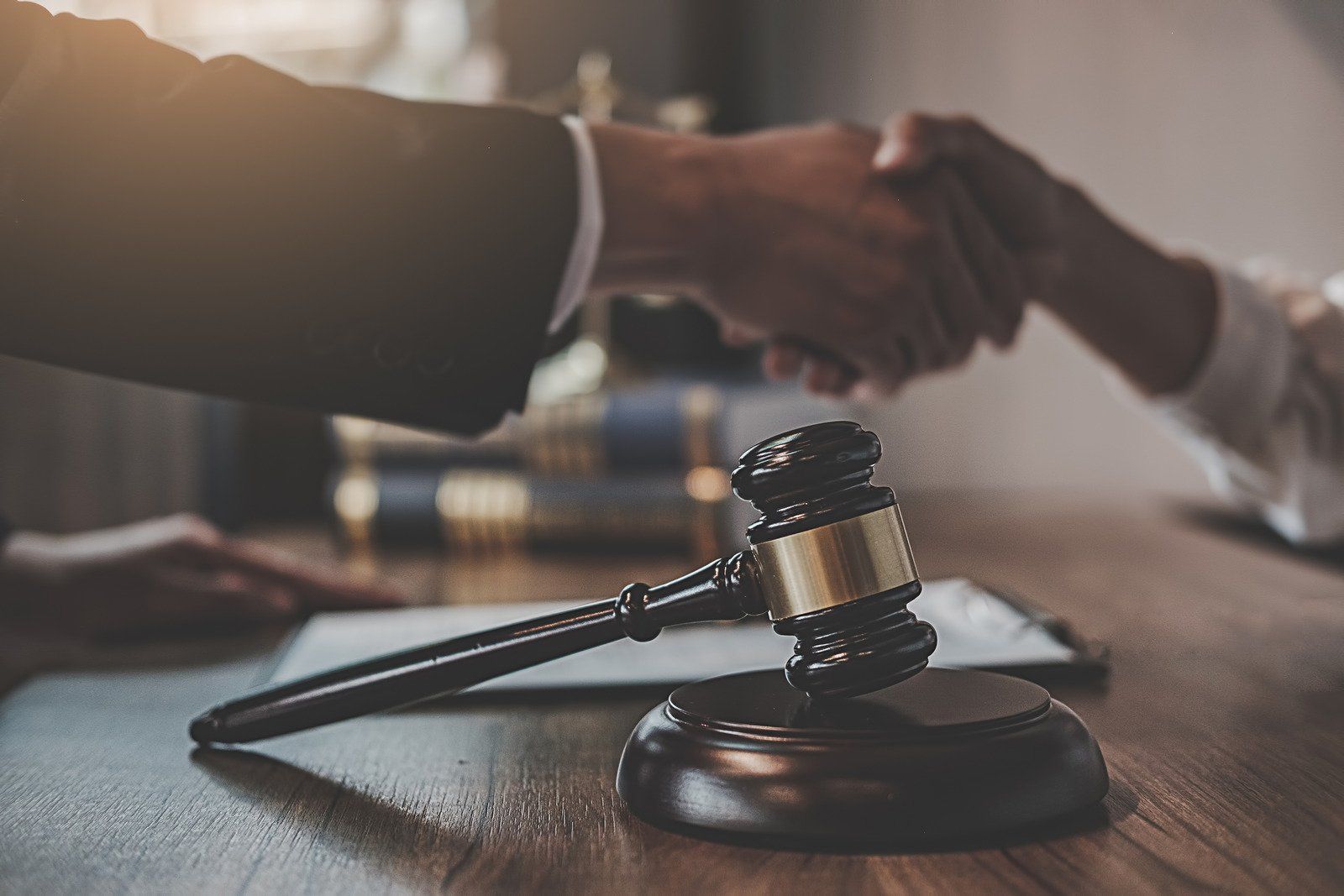Gavel on a wooden table with two people shaking hands in the background, likely a legal settlement.