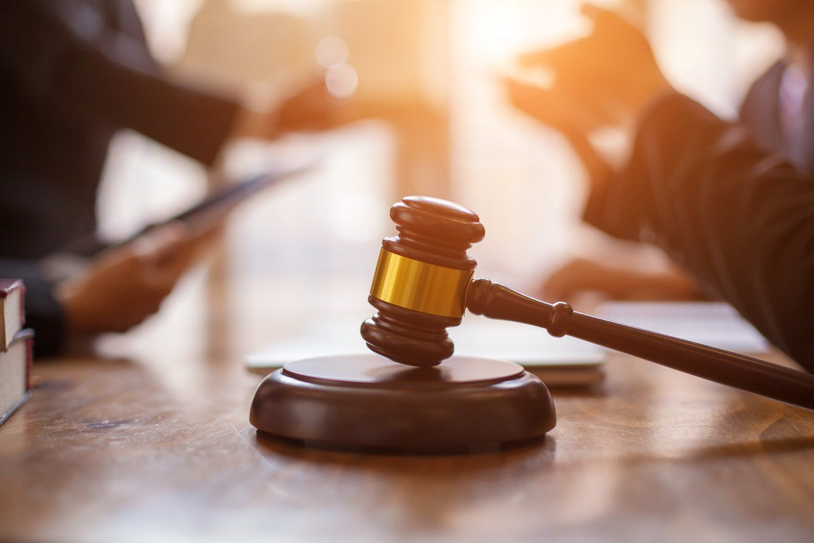 Gavel on wooden table with people in background, illuminated by sunlight.