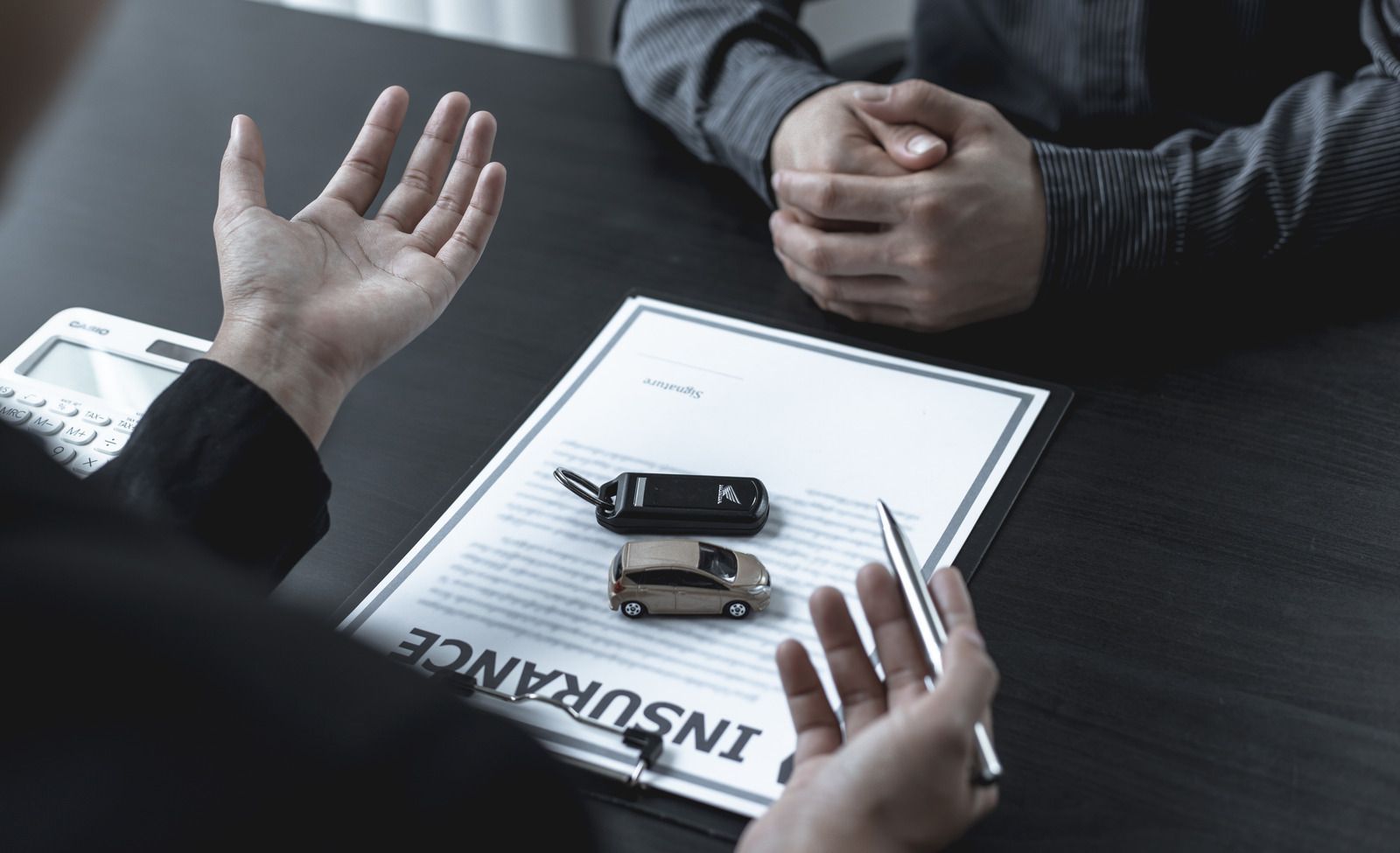 Hands gesturing over car keys and model car on an insurance document at a desk.