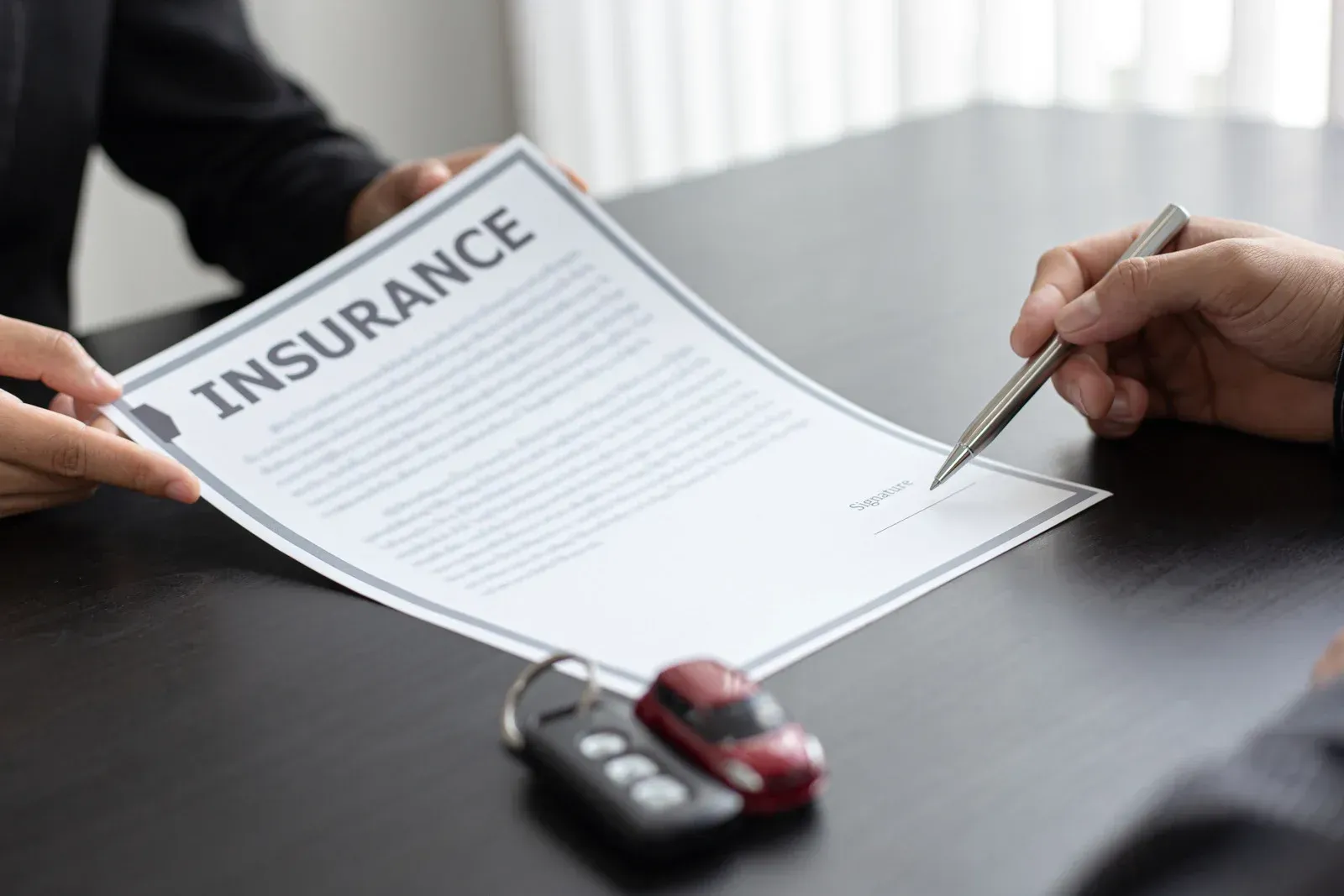 Person signing insurance document, car keys and toy car on table.