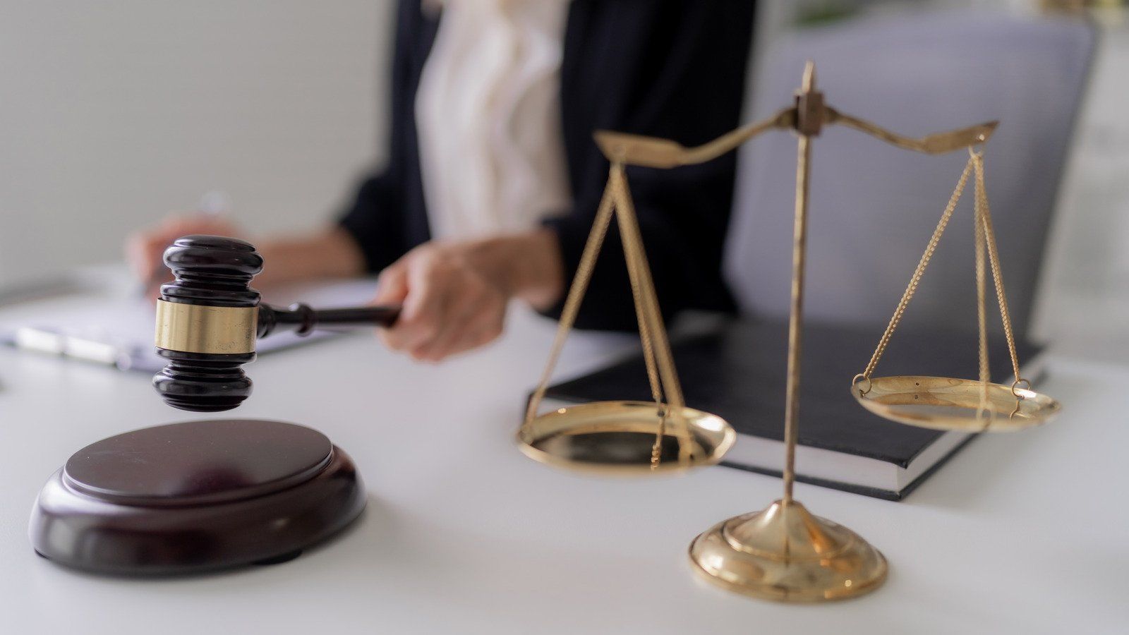 Gavel and scales of justice on a desk, a person in the background holds the gavel.