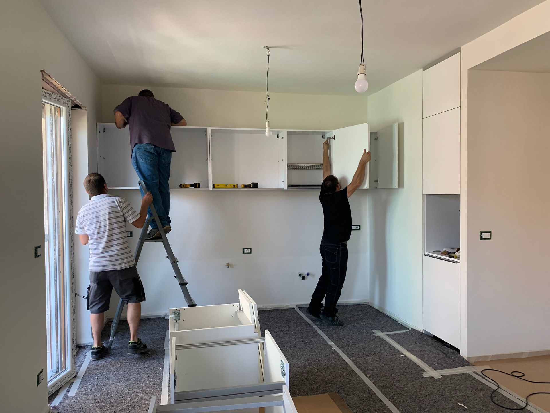 Bright white kitchen featuring custom cabinets with gold hardware and a large center island. Three people installing kitchen cabinets in an unfinished room with white walls and bare floor.