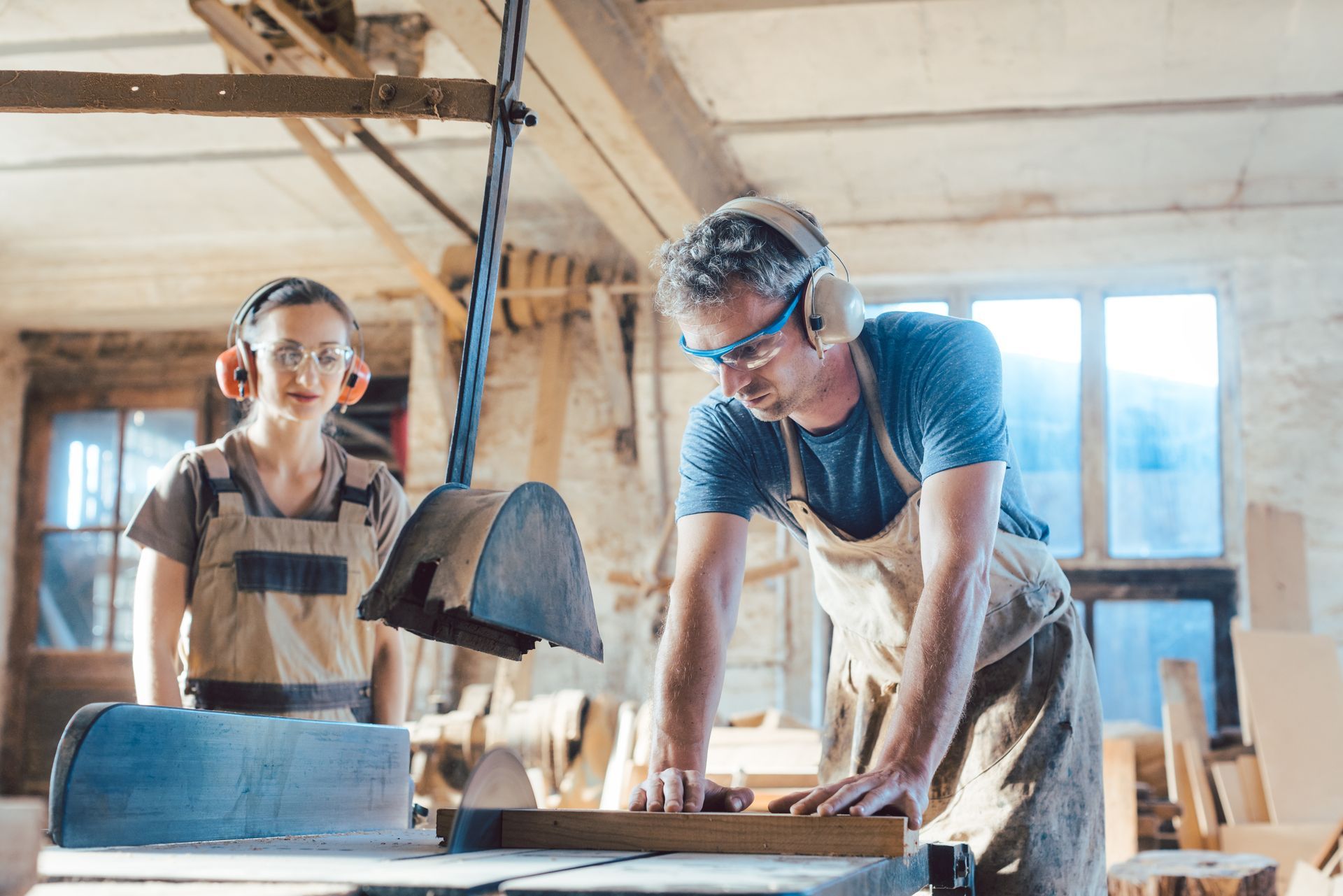 Two cabinet makers in ear protection cut a wood board on a table saw in a sunlit woodworking shop. Two cabinet makers in ear protection cut a wood board on a table saw in a sunlit woodworking shop.