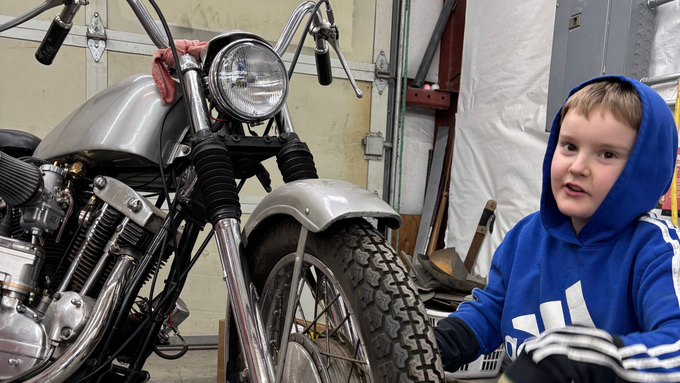 Blue-hooded child sitting beside a silver motorcycle in a garage, looking at the camera.