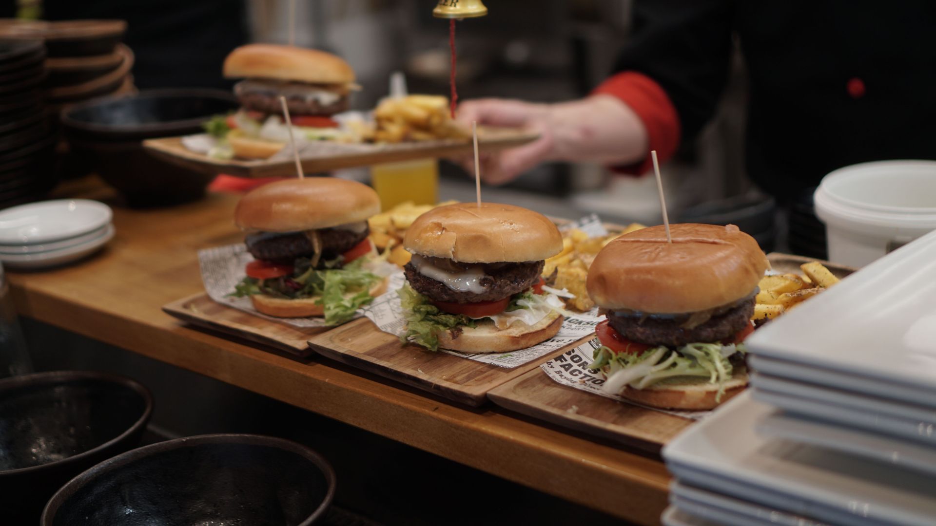Three gourmet burgers on a wooden serving board at a restaurant table, with stacked plates beside them.