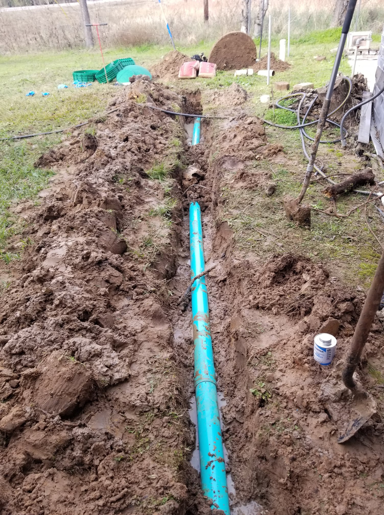 Trenched blue PVC pipe laid in muddy ground in a rural yard, with excavated soil on both sides