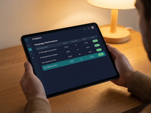 Hands holding a tablet displaying a dark-themed performance dashboard on a wooden desk next to a warm lamp.