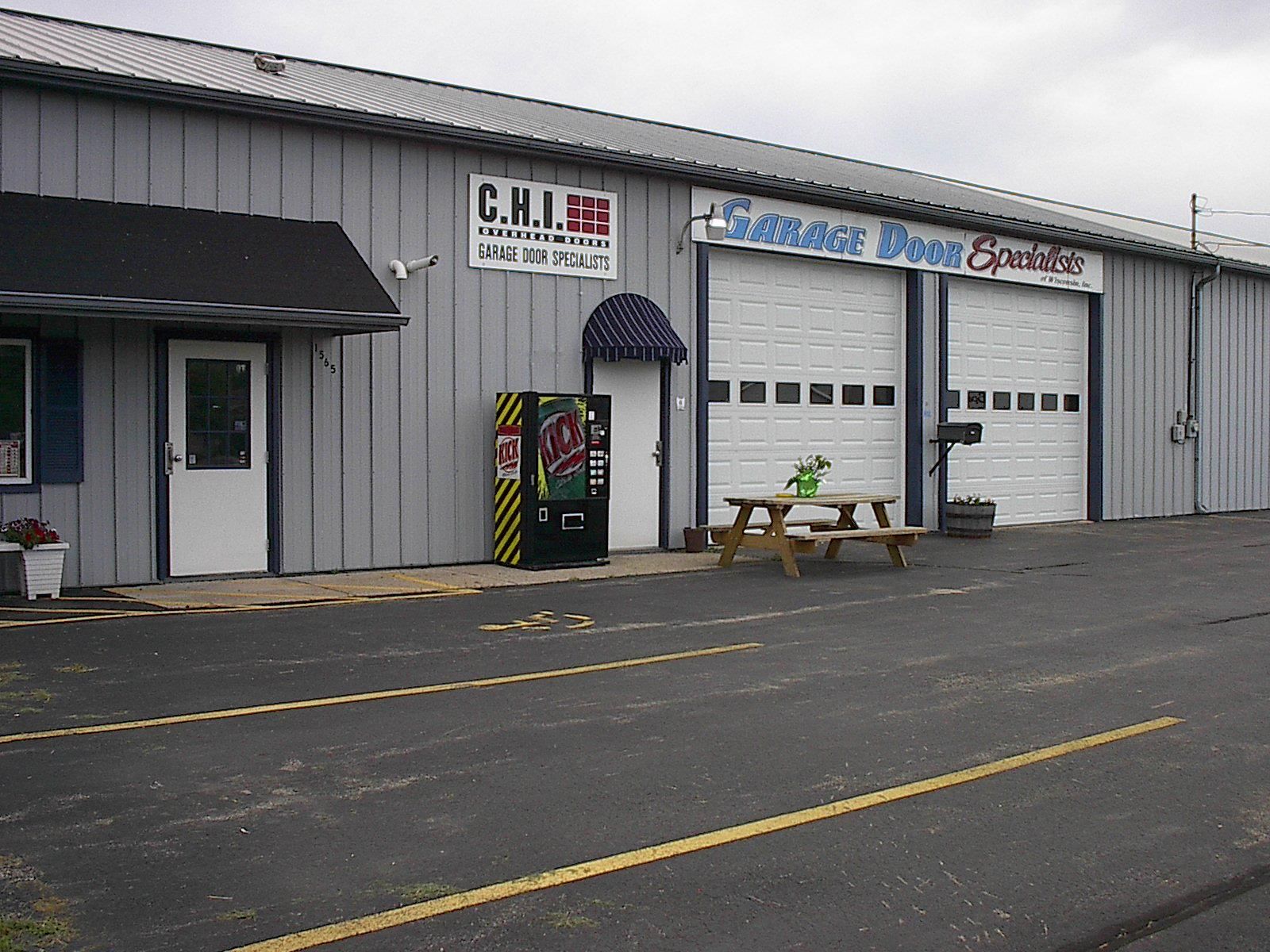 Gray storefront with a garage door, awning, vending machine, and picnic table in front.