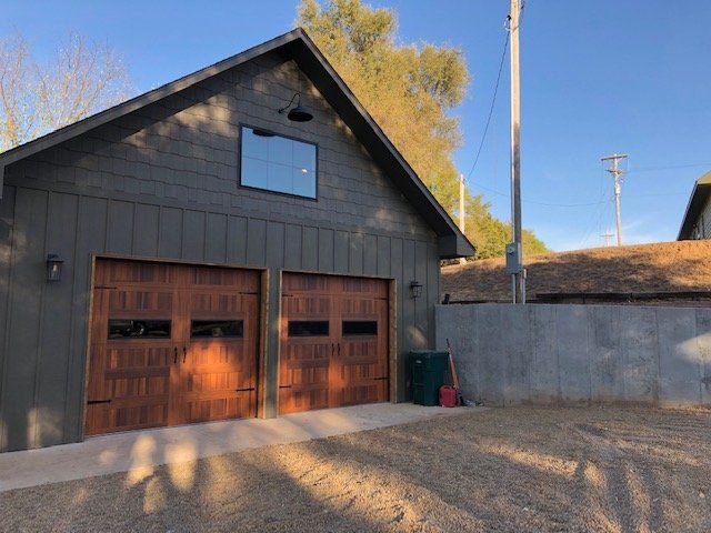 Dark gray garage with two wooden doors, gravel driveway, and trees under a blue sky.
