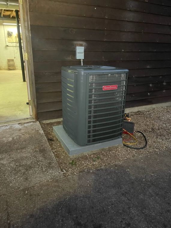 Outdoor air conditioner unit beside a brown-sided building, with a hose and concrete pad at dusk