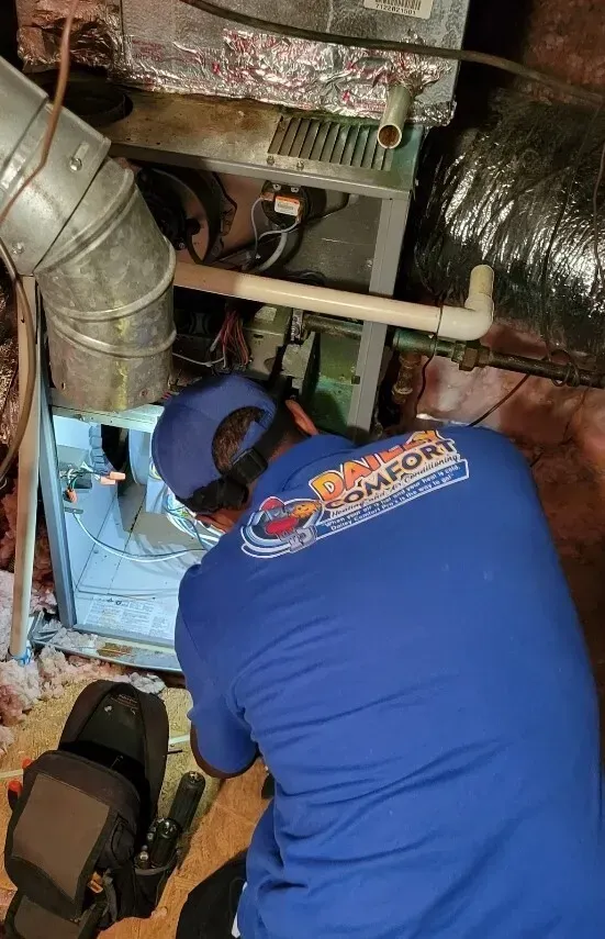 Technician in blue shirt works inside a crawlspace beneath a house, with ducts and insulation visible.