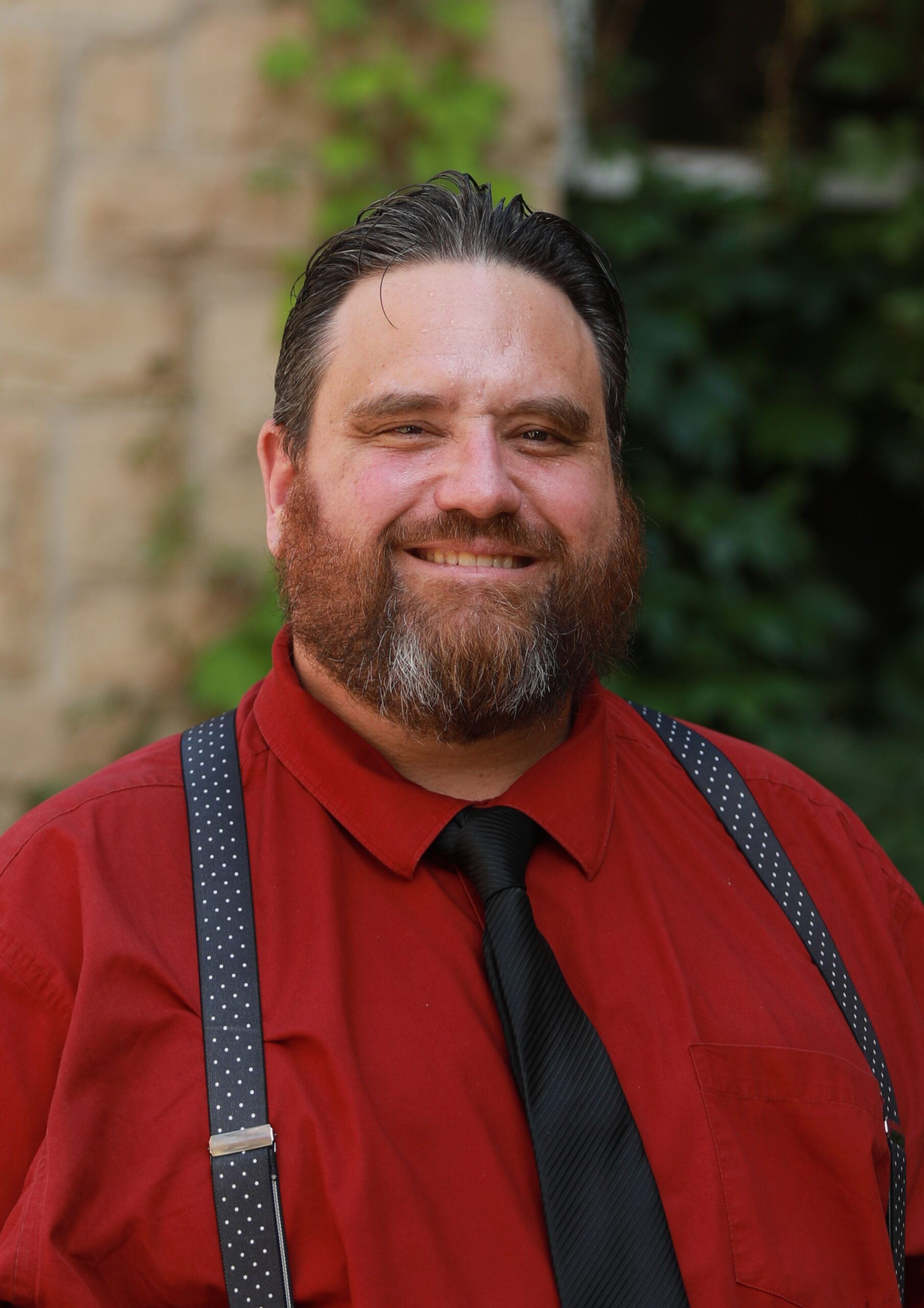 Smiling man in a red shirt, black tie, and suspenders standing outdoors.