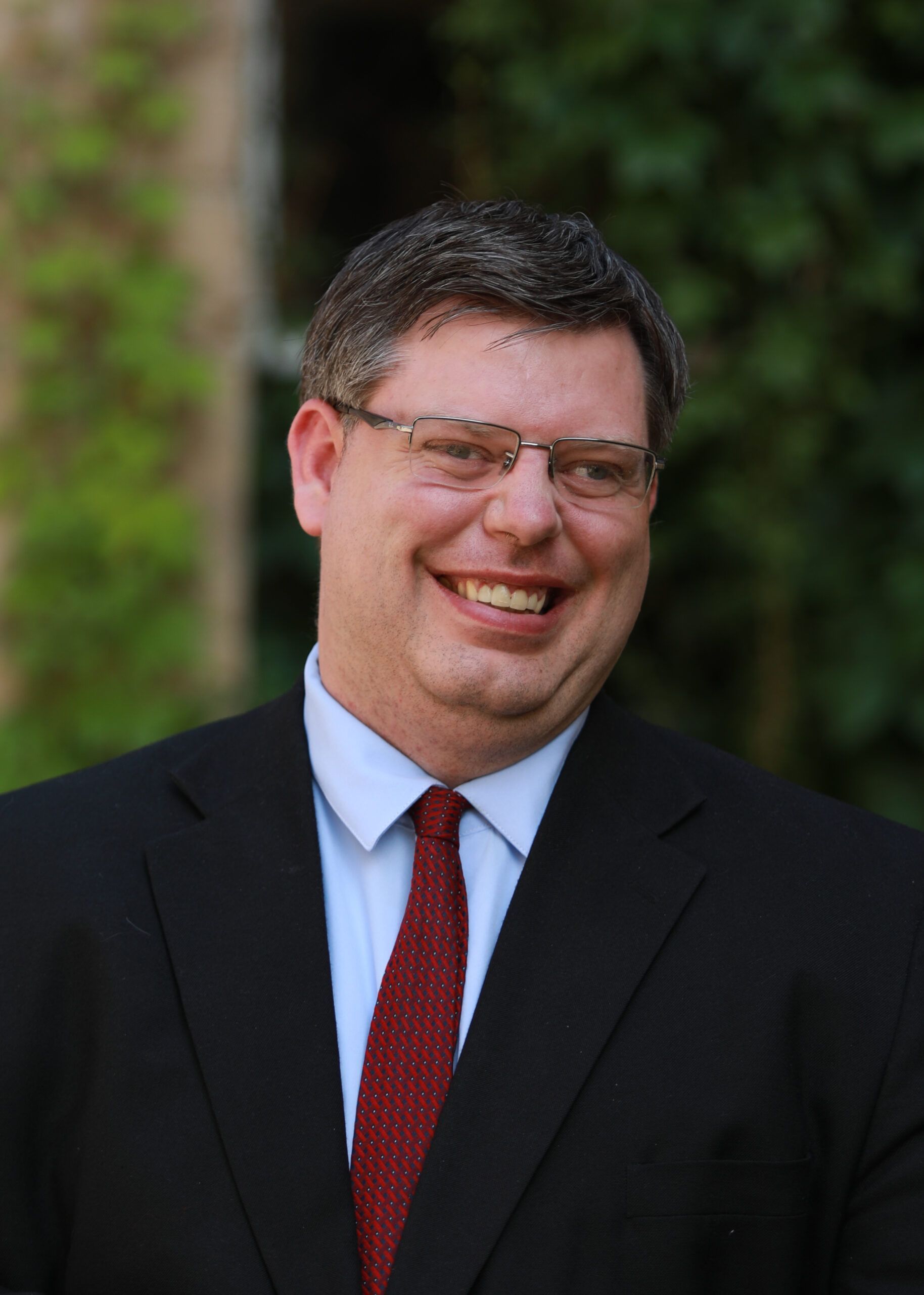 Smiling man in a dark suit and red tie outdoors with a blurred green background