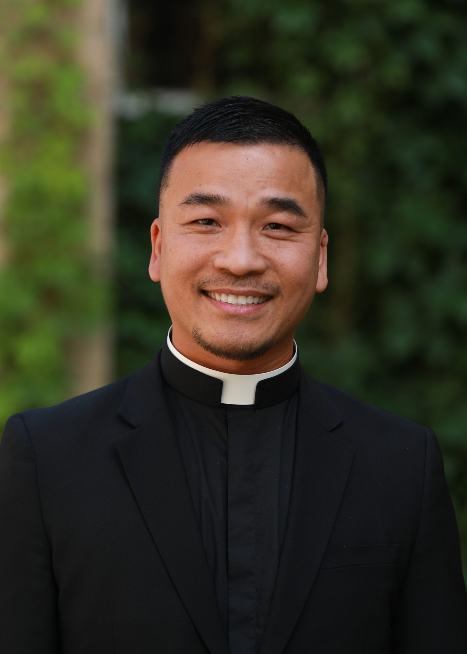 Smiling person in a black clerical suit with a white collar outdoors against a blurred green background