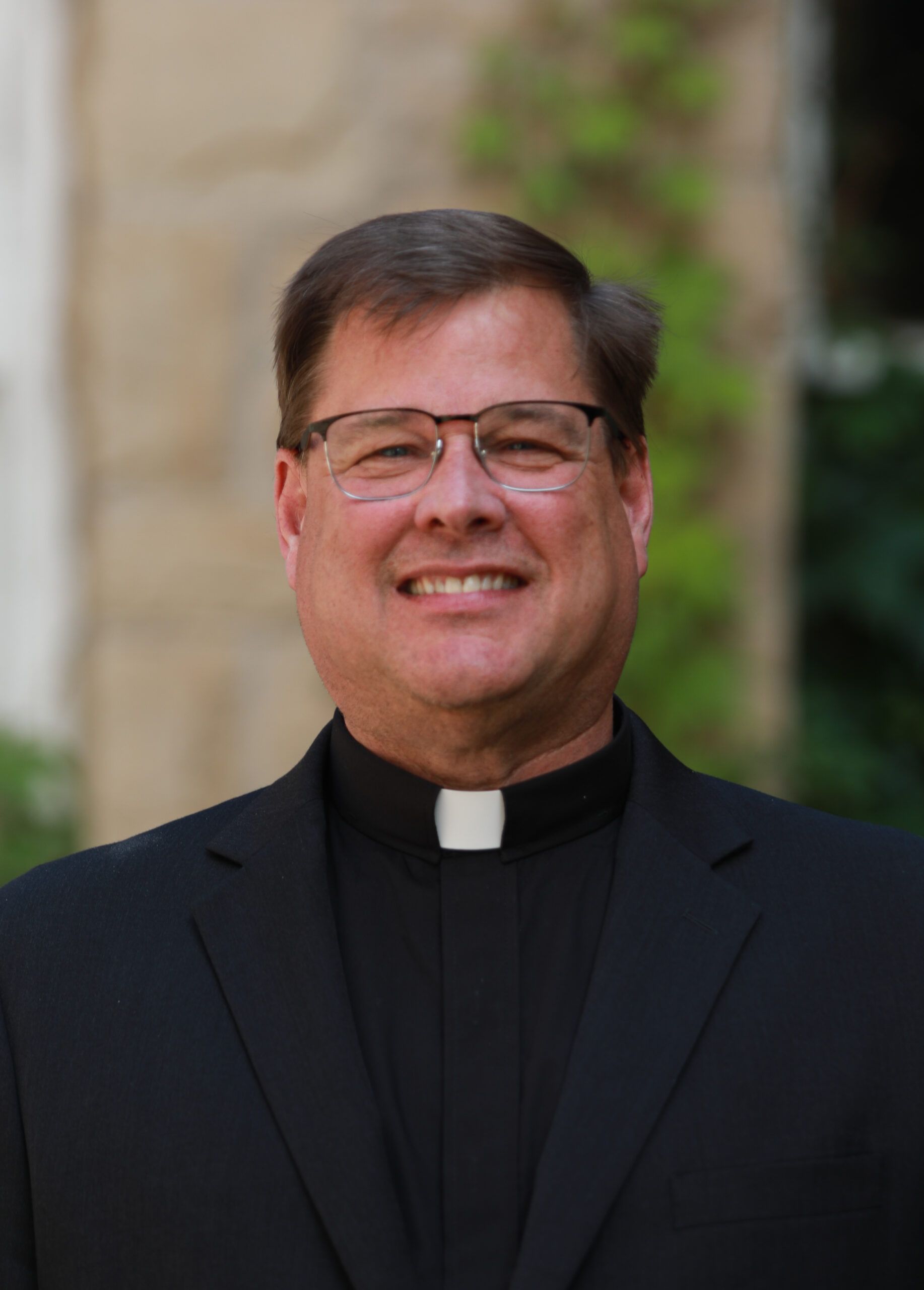 Man in a black clerical suit with a white collar smiling outdoors in front of a blurred stone background