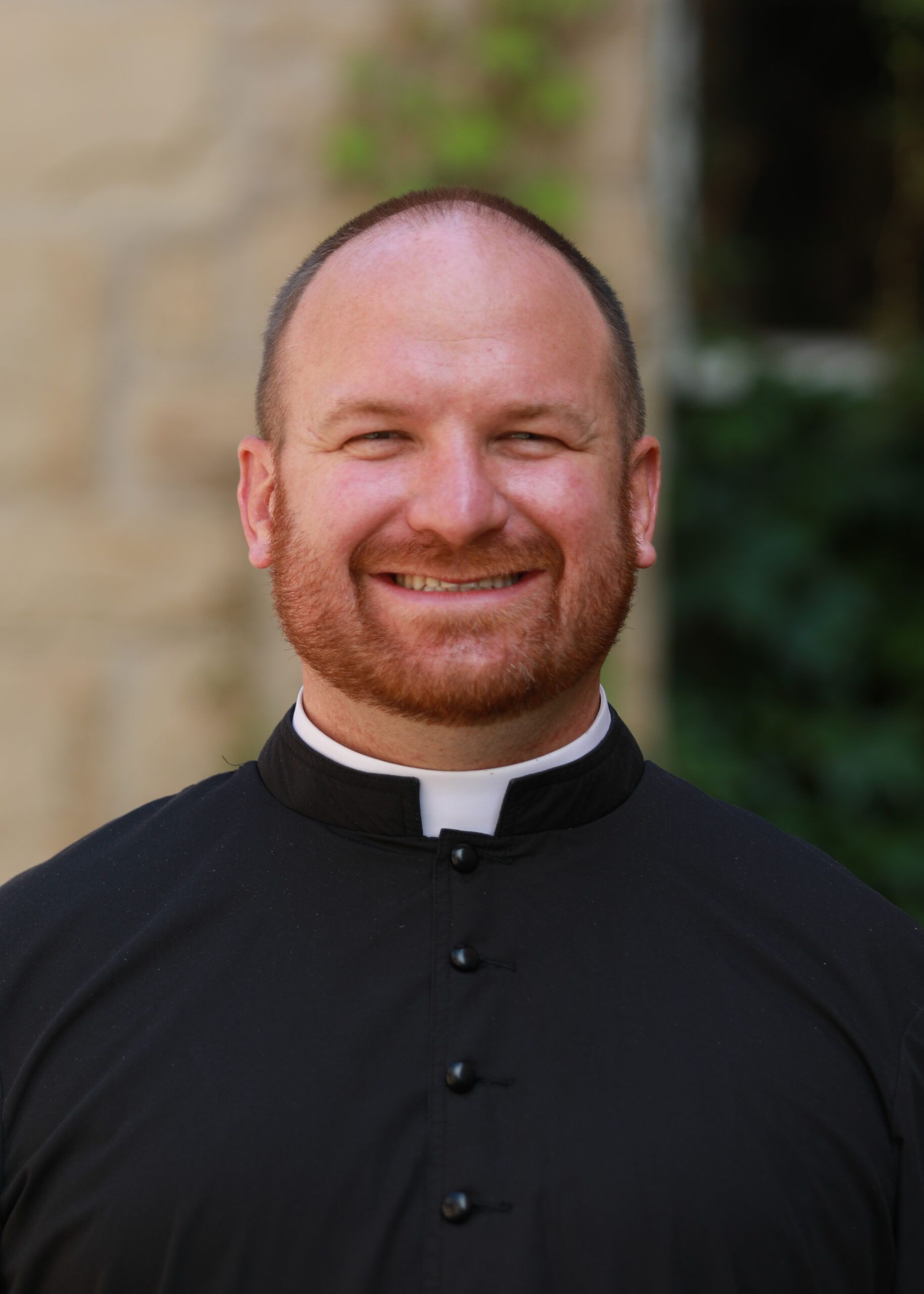 Smiling priest in a black clerical shirt with white collar, standing outdoors.