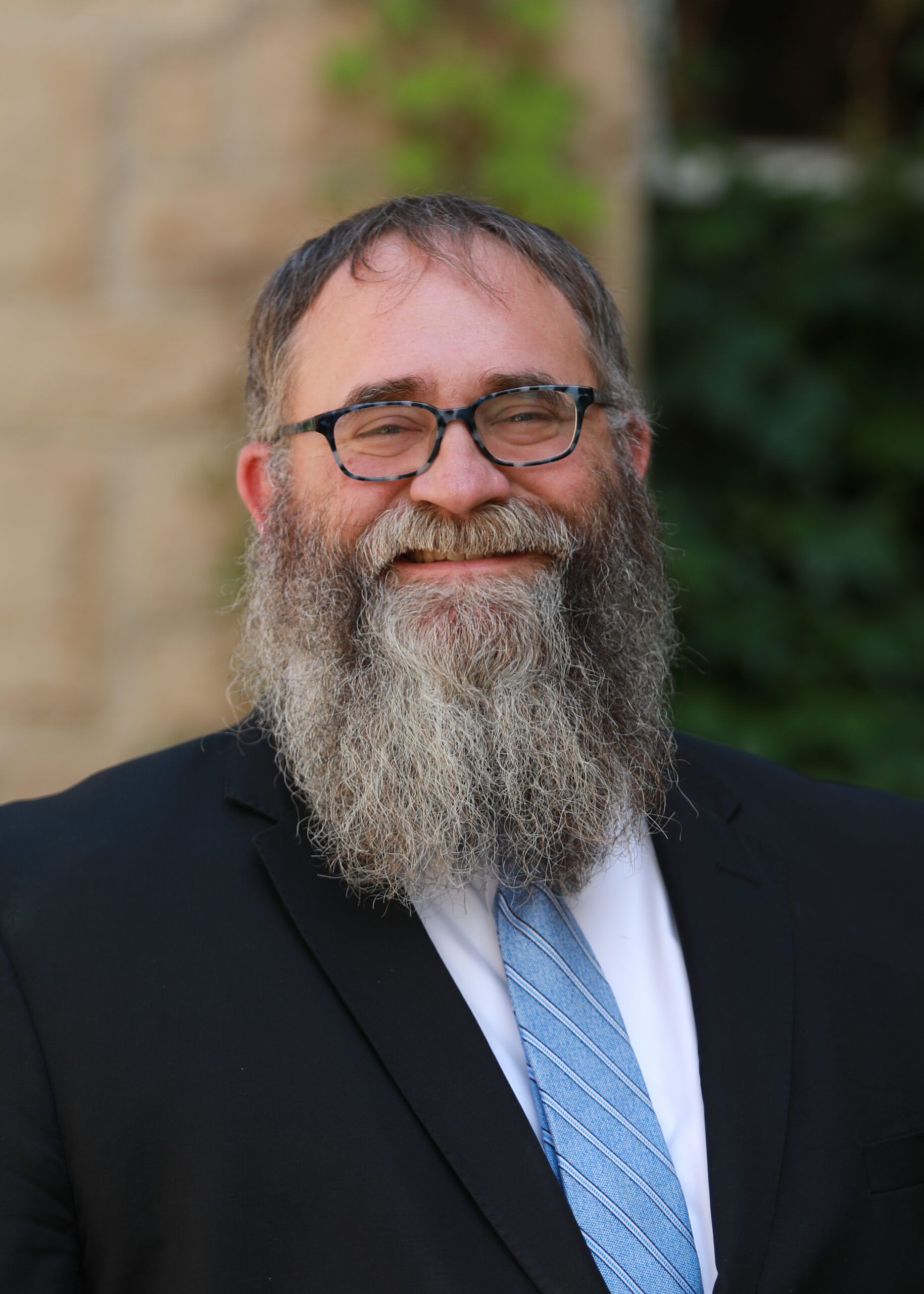 Smiling man in glasses with a long gray beard, wearing a dark suit and blue tie outdoors