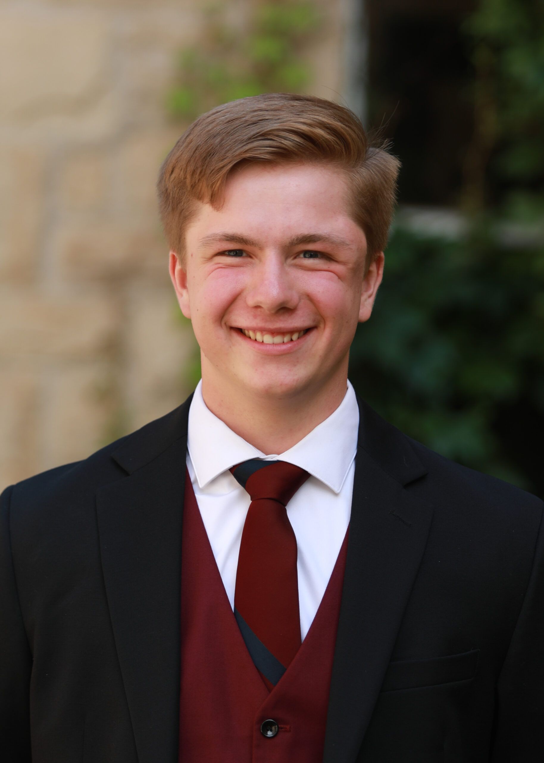 Young man in a black suit and burgundy vest smiling outdoors against a blurred background