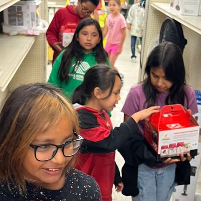 Children shop at a store, some holding red boxes, inspecting items on shelves.
