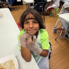 Girl with brown hair, smiling, wearing a hairnet, gloves, and a green shirt, leaning on a table.