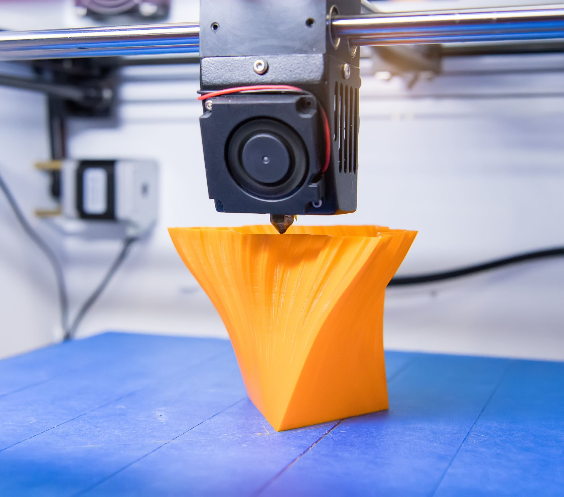 Ein 3D-Drucker extrudiert ein leuchtend orangefarbenes Objekt auf ein blaues Druckbett.