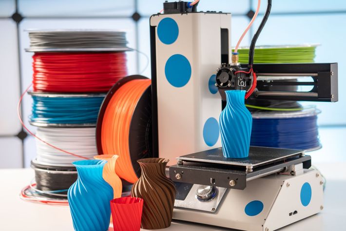 Ein 3D-Drucker druckt eine blaue Figur neben Spulen mit farbigem Filament.
