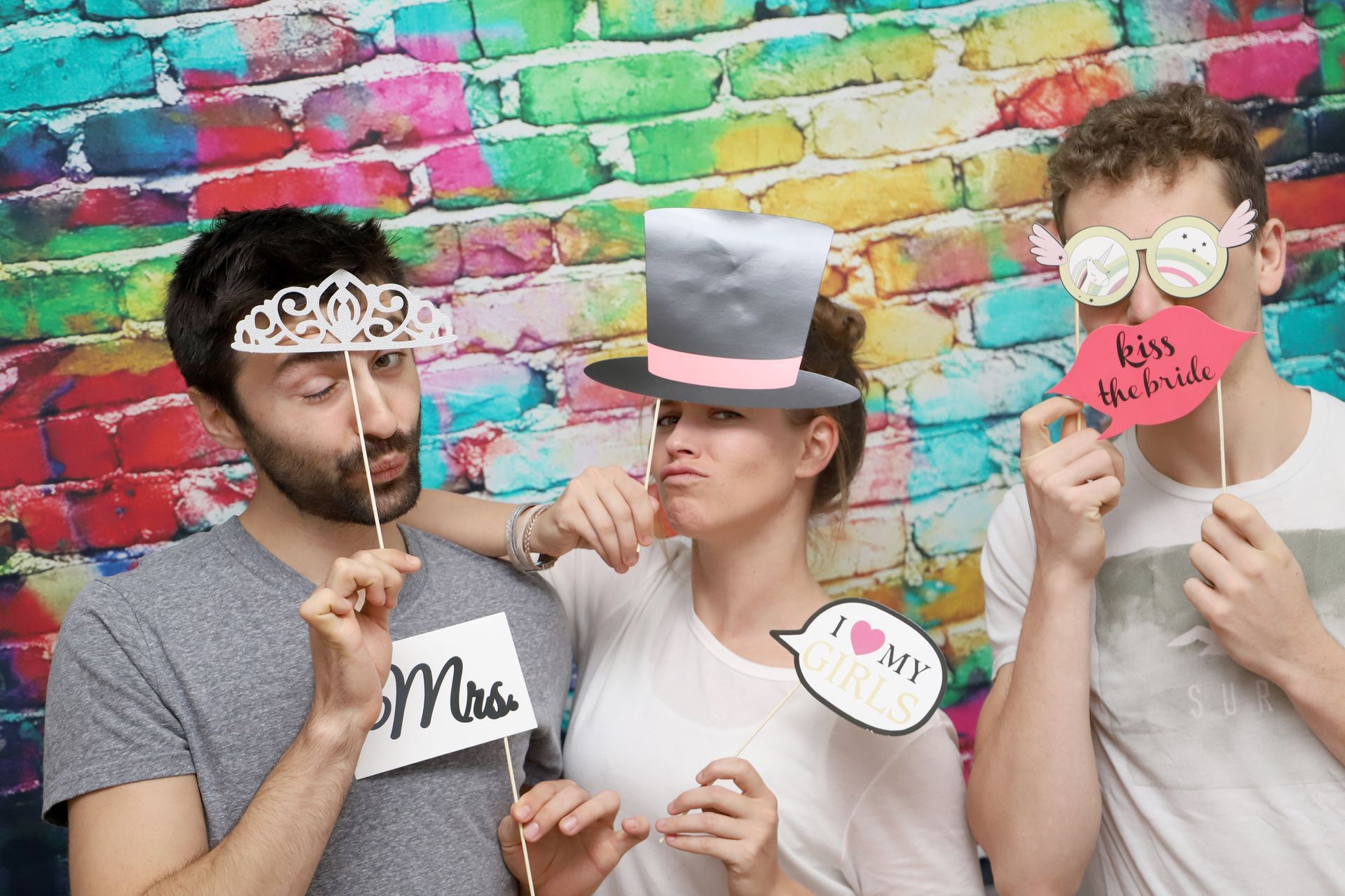 Drei Personen posieren mit Requisiten aus Papier vor einer farbenfrohen Graffitiwand in einer Fotobox.