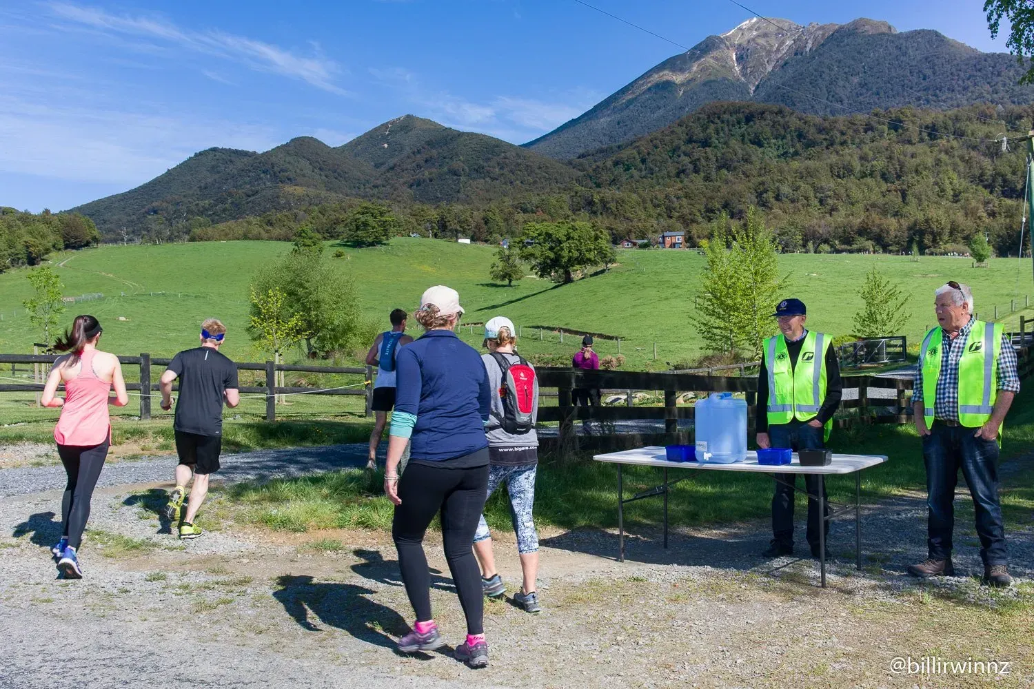 A group of people are standing around a table in a field with mountains in the background.